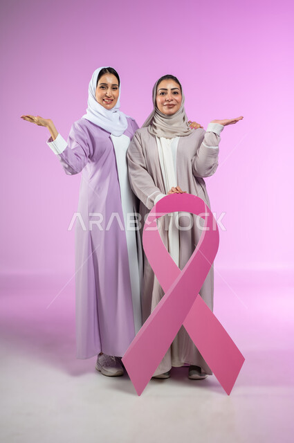 الامساك بشعار حملة التوعية، بورتريه لامراتان عربيتان خليجيتان سعوديتان محجبتان ترتديان العباءه الملونه، شعار الحملة العالمية لتحدي الأمراض الخبيثة، الاشارة بالكف إلى شيء ما و حمل عنصر معين، الجسم بالطول الكامل، خلفية ورديه، محتوى حساس، استخدام حساس