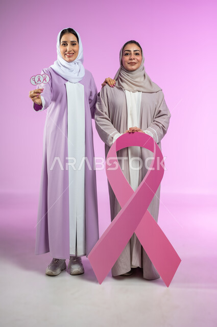 اليوم العالمي للتوعية والكشف المبكر عن سرطان الثدي, تحدي الأمراض الخبيثة, بورتريه لامراتان عربيتان خليجيتان سعوديتان محجبتان ترتديان العباءه الملونه، الامساك بشعار الحملة العالمية، الجسم بالطول الكامل، خلفية ورديه، محتوى حساس، استخدام حساس