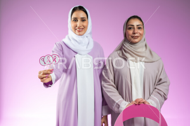 تحدي الأمراض الخبيثة, اليوم العالمي للتوعية والكشف المبكر عن سرطان الثدي، بورتريه مقرب لامراتان عربيتان خليجيتان سعوديتان محجبتان ترتديان العباءه الملونه، الامساك بشعار الحملة العالمية، خلفية ورديه، محتوى حساس، استخدام حساس
