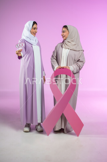 اليوم العالمي للتوعية والكشف المبكر عن سرطان الثدي, تحدي الأمراض الخبيثة, بورتريه لامراتان عربيتان خليجيتان سعوديتان محجبتان ترتديان العباءه الملونه، الامساك بشعار الحملة العالمية، الجسم بالطول الكامل، خلفية ورديه، محتوى حساس، استخدام حساس