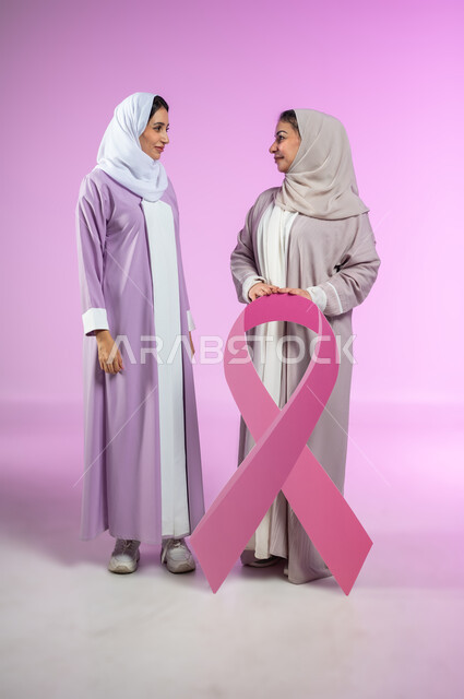 تحدي الأمراض الخبيثة, اليوم العالمي للكشف المبكر عن سرطان الثدي, بورتريه لامراتان عربيتان خليجيتان سعوديتان محجبتان ترتديان العباءه الملونه تمسكان  بشعار الحملة العالمية، الجسم بالطول الكامل، خلفية ورديه، محتوى حساس، استخدام حساس