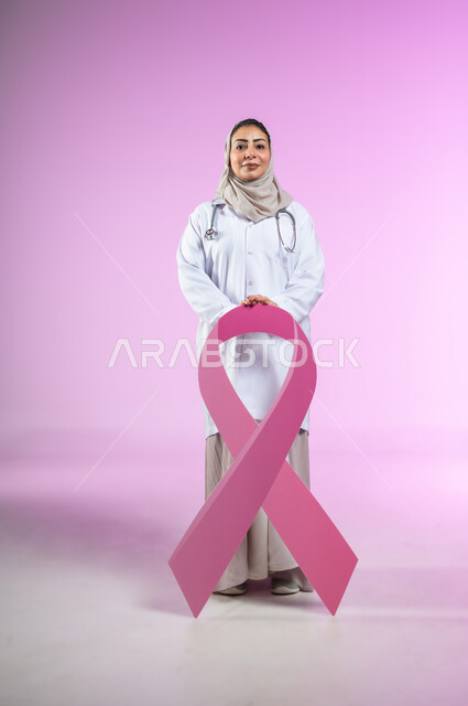 النظر الى الكاميرا بإيماءات السرور، اليوم العالمي للتوعية والكشف المبكر عن سرطان الثدي, تحدي الأمراض الخبيثة, بورتريه لإمرأة عربية خليجية سعودية ترتدي العباءة واللباس الطبي و السماعة تمسك بيديها شعار الحملة العالمية, الجسم بالطول الكامل, خلفية ورديه, محتوى حساس, استخدام حساس