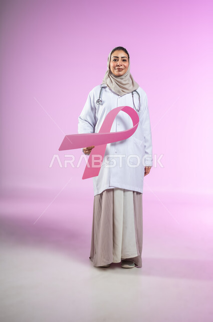 اليوم العالمي للتوعية والكشف المبكر عن سرطان الثدي, تحدي الأمراض الخبيثة,  بورتريه لإمرأة عربية خليجية سعودية مبتسمة ترتدي العباءة واللباس الطبي و السماعة تمسك بيدها شعار الحملة العالمية, الجسم بالطول الكامل, خلفية ورديه, محتوى حساس, استخدام حساس