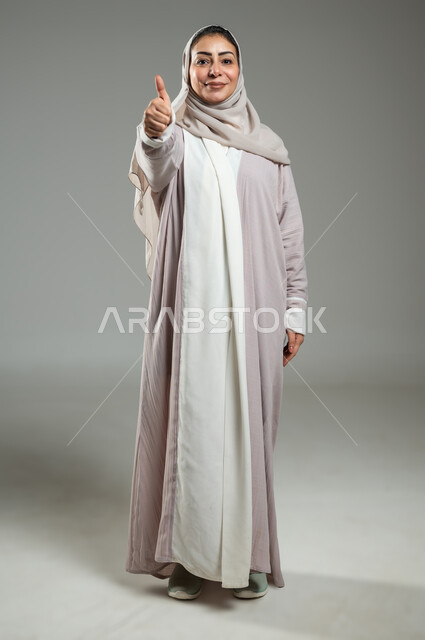 رفع اصبع الابهام للاعلى بإيماءات الرضا والسرور، بورتريه لامرأة عربية خليجية سعودية مبتسمة محجبة ترتدي العباءة الملونة تنظر إلى الكاميرا، اليوم العالمي للتوعية والكشف المبكر عن سرطان الثدي، الجسم بالطول الكامل، خلفية رمادية، محتوى حساس، استخدام حساس