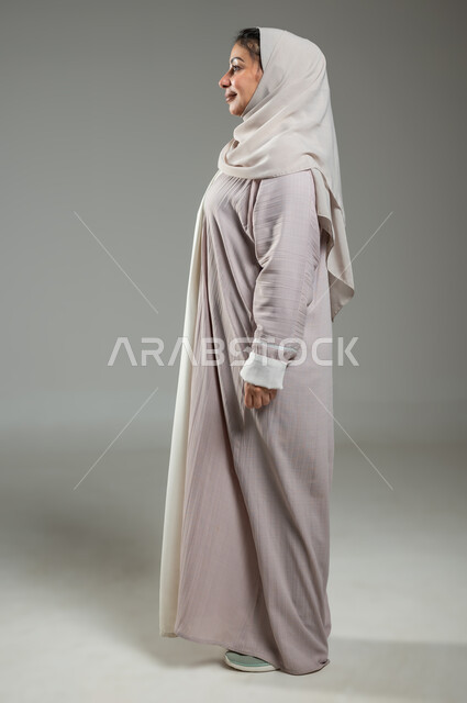 التزيين و التجمل والاهتمام بالمظهرالخارجي، بورتريه من الجانب لإمراة عربية سعودية خليجية ترتدي العباءة والحجاب تقف باستقامة بإيماءات الاعتزاز بالنفس و الانضباط، ايماءات وتعابير الثقة، خلفية رمادية، محتوى حساس، استخدام حساس