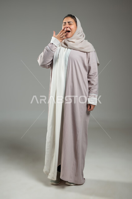 السهر لوقت متأخر، الحاجة للنوم والراحة، بورتريه لإمرأه عربية خليجية سعودية محجبة ترتدي عباءة تقوم بالتثاؤب، إيماءات النعاس والتعب، تعابير الكسل والخمول، صورة الجسم بالطول الكامل، خلفية رمادية، محتوى حساس، استخدام حساس