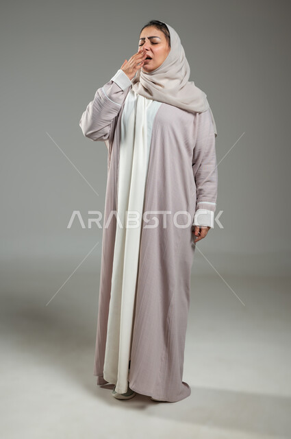 الحاجة للنوم والراحة، بورتريه لإمرأه عربية خليجية سعودية محجبة ترتدي عباءة تقوم بالتثاؤب، إيماءات النعاس والتعب، تعابير الكسل والخمول، السهر لوقت متأخر، صورة الجسم بالطول الكامل، خلفية رمادية، محتوى حساس، استخدام حساس