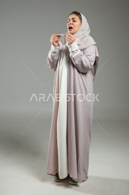 استعمال المناديل الورقيه أثناء العطس، الشعور بالتعب والمرض، الإصابة بالزكام والانفلونزا, تعابير تدل على العطاس والإرهاق، بورتريه لإمرأة عربية سعودية خليجية محجبة ترتدي العباءة تعاني من الحساسية, الجسم بالطول الكامل, خلفية رمادية، محتوى حساس، استخدام حساس