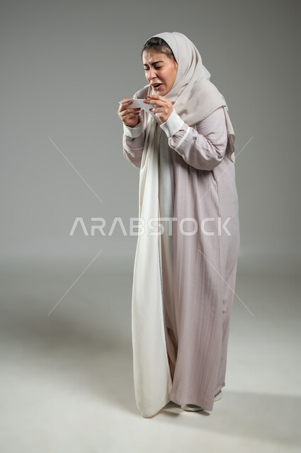 Suffering from cold and flu, expressions indicating sneezing and fatigue, using tissues while sneezing, feeling tired and sick, portrait of a veiled Saudi Arabian Gulf woman wearing an abaya suffering from allergies, full-length body, gray background, sensitive content, sensitive use