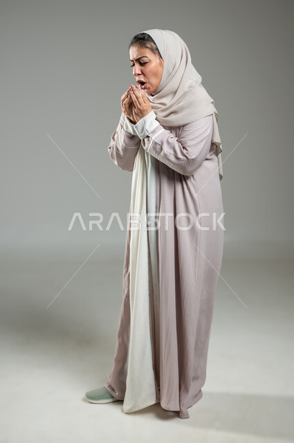 الشعور بالمرض والكحة، بورتريه لامرأه عربية خليجية سعودية ترتدي العباءة والحجاب تعاني من السعال، الانفلونزا والتهاب الرئه، الاصابة بفايروسات صدرية، الجسم بالطول الكامل، خلفية رمادية، محتوى حساس، استخدام حساس
