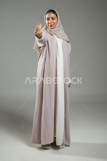 تعابير طلب الإنتظار والتمهل، مفهوم الصبر والتريث، بورتريه لامرأة عربية خليجية سعودية محجبة ترتدي عباءة وردية ترفع يدها بتعابير تدل على التوعد والتهديد لشخص ما، صورة الجسم بالطول الكامل، خلفية رمادية، محتوى حساس، استخدام حساس