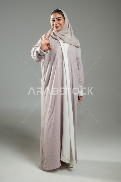 التعبير عن الجودة والاتقان، القبول والموافقة والرضا، بورتريه لإمراة عربية سعودية خليجية ترتدي العباءة والحجاب تشير بيدها بإيماءات وتعابير وجه و يدين تدل على الاعجاب، علامة الجودة الامتياز والتميز، الجسم بالطول الكامل، خلفية رمادية، محتوى حساس، استخدام حساس
