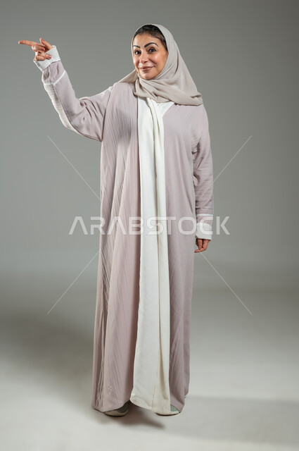 إشارات وحركات يدين تدل على هذا المكان (هنا)، تحديد موقع شيء معين، بورتريه لامرأة عربية خليجية سعودية محجبة ترتدي عباءة وردية تشير بإصبع السبابة إلى جهة اليمين، صورة الجسم بالكامل، خلفية رمادية، محتوى حساس، استخدام حساس