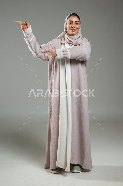 تحديد موقع شيء معين، إشارات وحركات يدين تدل على هذا المكان (هنا)، بورتريه لامرأة عربية خليجية سعودية محجبة ترتدي عباءة وردية تشير بإصبع السبابة إلى جهة اليمين، صورة الجسم بالكامل، خلفية رمادية، محتوى حساس، استخدام حساس
