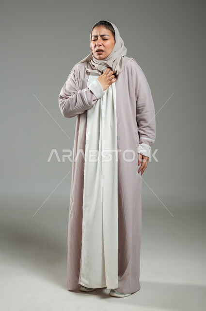 Gestures of fatigue, exhaustion, and psychological pressures, difficult health problems, a portrait of a veiled Saudi Gulf Arab woman wearing an abaya, placing her hand on her heart, feeling a bad state of tingling and pain, gray background, sensitive content, sensitive use