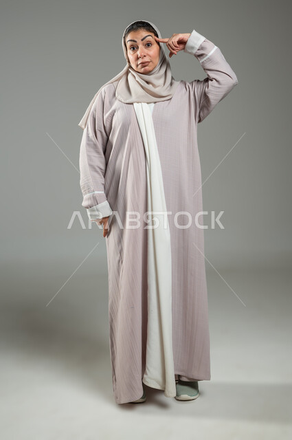 Contemplating, concentrating and planning for the future, trying to find a solution to a problem, portrait of a veiled Saudi Arabian Gulf woman wearing an abaya, placing her hand on her head and looking at the camera with gestures of thought and contemplation, full-length body, gray background, sensitive use, sensitive content