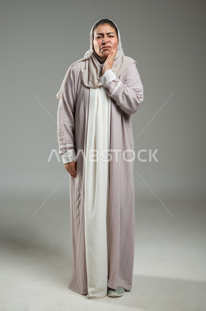 الشعور بألم الأسنان وتسوس الأضراس, الحاجه لزيارة طبيب الأسنان، بورتريه لإمراة عربية سعودية خليجية ترتدي العباءة والحجاب تضع يدها على خدها، إيماءات وجه ويدين تدل على الالم ووجع الفم، الجسم بالطول الكامل، خلفية رمادية، استخدام حساس، محتوى حساس