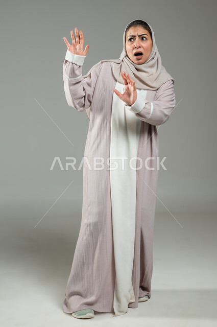 الذعر والرعب والرهبة من شيء ما، حماية النفس من ضرر خارجي, بورترية لإمراة عربية سعودية خليجية ترتدي العباءة والحجاب ترفع كفيها لأعلى بإيماءات الخوف و طلب التوقف والإمتناع، الجسم بالطول الكامل، خلفية رمادية، استخدام حساس، محتوى حساس