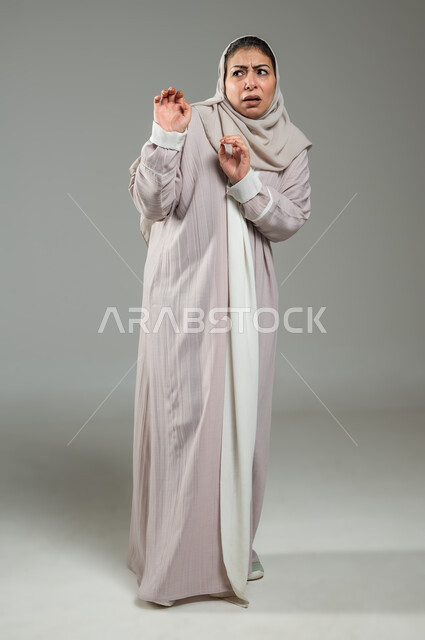 الرعب والذعر والرهبة من شيء ما، حماية النفس من ضرر خارجي, بورترية لإمراة عربية سعودية خليجية ترتدي العباءة والحجاب ترفع كفيها لأعلى بإيماءات الخوف و طلب التوقف والإمتناع، الجسم بالطول الكامل، خلفية رمادية، استخدام حساس، محتوى حساس