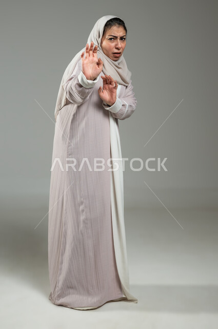 حماية النفس من ضرر خارجي, الرعب والذعر والرهبة من شيء ما، بورترية لإمراة عربية سعودية خليجية ترتدي العباءة والحجاب ترفع كفيها لأعلى بإيماءات الخوف و طلب التوقف والإمتناع، الجسم بالطول الكامل، خلفية رمادية، استخدام حساس، محتوى حساس