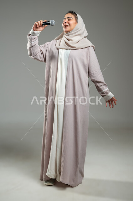 قضاء وقت ممتع بأداء العروض الصوتية، بورتريه لإمرأة عربية سعودية خليجية محجبة ترتدي العباءة تمسك بيدها المايكروفون بإيماءات السرور، مفهوم الفن وتأثيره الايجابي، ممارسة موهبة الغناء، الجسم بالطول الكامل، خلفية رمادية، استخدام حساس، محتوى حساس
