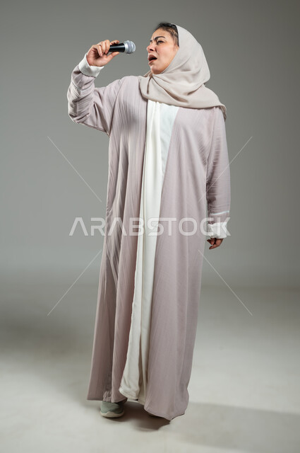 Spending a good time performing vocal performances, portrait of a veiled Saudi Arabian Gulf woman wearing an abaya, holding the microphone in her hand with gestures of pleasure, the concept of art and its positive effect, practicing the talent of singing, full-length body, gray background, sensitive use, sensitive content