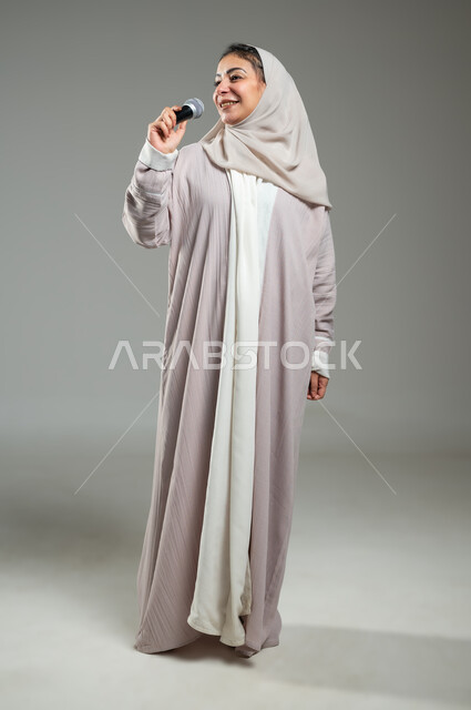ممارسة موهبة الغناء، قضاء وقت ممتع بأداء العروض الصوتية، بورتريه لإمرأة عربية سعودية خليجية محجبة ترتدي العباءة تمسك بيدها المايكروفون بإيماءات السرور، مفهوم الفن وتأثيره الايجابي، الجسم بالطول الكامل، خلفية رمادية، استخدام حساس، محتوى حساس