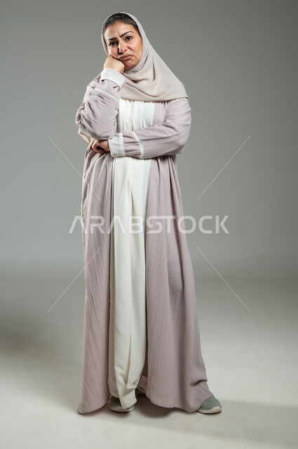 الشعور بالجهل وعدم المعرفة، بورتريه لإمرأة عربية سعودية خليجية محجبة ترتدي العباءة تضع يدها على ذقنها تنظر إلى الكاميرا بإيماءات الشعور والحيرة، التردد بالثقة بخصوص أمر معين، الجسم بالطول الكامل، خلفية رمادية، استخدام حساس، محتوى حساس