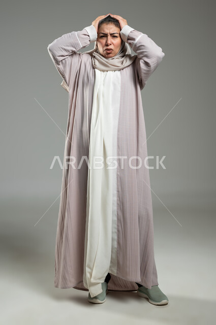 الحاجة للهدوء والراحة، الإحساس بألم الرأس والصداع، المعاناة من آلام الشقيقة، بورتريه لإمرأة عربية خليجية سعودية محجبة ترتدي عباءة وردية تضع يديها على رأسها بايماءات التعب والارهاق، صورة الجسم بالطول الكامل، خلفية رمادية، محتوى حساس، استخدام حساس