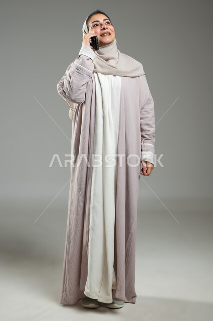 قضاء اوقات ممتعة بتبادل الاخبار والأحاديث مع الاصدقاء والأهل، بورتريه لإمرأة عربية سعودية خليجية محجبة ترتدي العباءة تقوم بالتحدث عبر الهاتف المحمول، ايماءات السعادة و السرور بإجراء مكالمة هاتفية، الجسم بالطول الكامل، خلفية رمادية، استخدام حساس، محتوى حساس