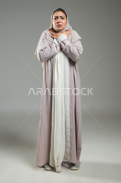 ايماءات الاختناق وعدم التنفس، تعابير الخوف والحزن، بورتريه لإمراة عربية سعودية خليجية ترتدي العباءة والحجاب تضع يدها على رقبتها بايماءات وجه ويدين تدل على الشعور بالضيق ، الجسم بالطول الكامل، خلفية رمادية، استخدام حساس، محتوى حساس