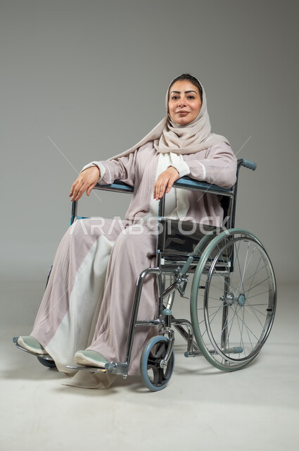 تأمين معدات للمساعده على الحركة والتنقل, تحسين حياة ذوي الاعاقه الحركيه، ايماءات السعاده و التفاؤل بالحياه, بورتريه لإمرأة عربية سعودية خليجية مبتسمة ترتدي العباءة والحجاب تجلس على كرسي متحرك، خلفية رمادية، استخدام حساس، محتوى حساس