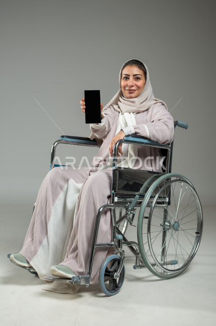 العنايه بأصحاب الهمم وتأمين احتياجاتهم الخاصة، عرض شاشة سوداء فارغه على الهاتف المحمول, بورتريه لإمرأة عربية سعودية خليجية محجبة مبتسمة ترتدي العباءة تجلس على كرسي متحرك، تسهيل حياة المعاقين باستخدام التقنيه والتكنولوجيا, الجسم بالطول الكامل، حلفية رمادية