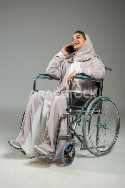تسهيل حياة ذوي الهمم باستخدام التقنيه والتكنولوجيا، التواصل مع الأهل والأصدقاء، بورتريه لإمرأة عربية سعودية خليجية محجبة مبتسمة ترتدي العباءة، تجلس على كرسي متحرك تمسك الهاتف المحمول تقوم بإجراء مكالمة هاتفية، الجسم بالطول الكامل، خلفية رمادية