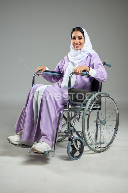 تحسين حياة ذوي الاعاقه الحركيه، تأمين معدات للمساعده على الحركة والتنقل, ايماءات السعاده و التفاؤل بالحياه, بورتريه لإمرأة عربية سعودية خليجية مبتسمة ترتدي العباءة والحجاب تجلس على كرسي متحرك، خلفية رمادية، استخدام حساس، محتوى حساس