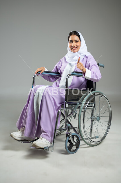 تعابير وإيماءات تدل على الإعجاب والرضى, بورتريه لإمرأة عربية سعودية خليجية محجبة مبتسمة ترتدي العباءة تجلس على كرسي متحرك, أجهزة ومعدات تساعد ذوي الهمم على الحركة والتنقل، علامات الجودة والتميز، خلفية رمادية، استخدام حساس، محتوى حساس