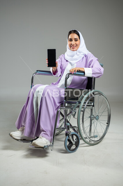 عرض شاشة سوداء فارغه على الهاتف المحمول, بورتريه لإمرأة عربية سعودية خليجية محجبة مبتسمة ترتدي العباءة تجلس على كرسي متحرك, تسهيل حياة المعاقين باستخدام التقنيه والتكنولوجيا، حلفية رمادية، استخدام حساس، محتوى حساس