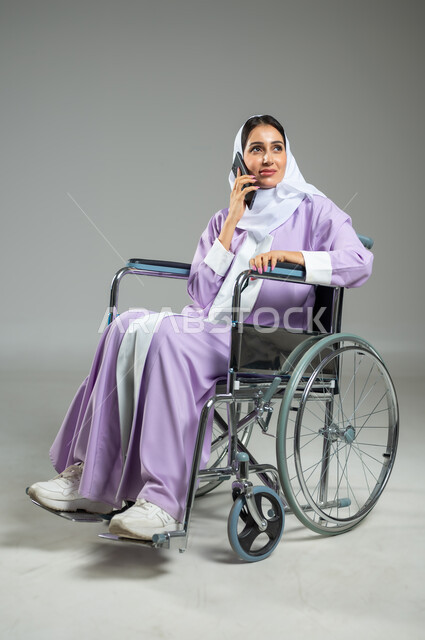 التواصل مع الأهل والأصدقاء، تسهيل حياة ذوي الهمم باستخدام التقنيه والتكنولوجيا، بورتريه لإمرأة عربية سعودية خليجية محجبة مبتسمة ترتدي العباءة، تجلس على كرسي متحرك تمسك الهاتف المحمول تقوم بإجراء مكالمة هاتفية، خلفية رمادية، استخدام حساس، محتوى حساس