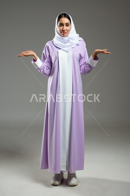 الشعور بالحيرة و التردد بخصوص أمر معين، بورتريه لإمرأة عربية سعودية خليجية محجبة ترتدي العباءة ترفع كفات يدها لأعلى بإيماءات و تعابير وجه ويدين تدل على عدم المعرفة أمر ما، الجسم بالطول الكامل، خلفية رمادية، استخدام حساس، محتوى حساس
