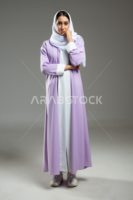 Feeling of ignorance and lack of knowledge, portrait of a veiled Saudi Arabian Gulf woman wearing an abaya, placing her hand on her chin, looking at the camera with gestures of feeling and bewilderment, hesitation with confidence regarding a certain matter, full-length body, gray background, sensitive use, sensitive content