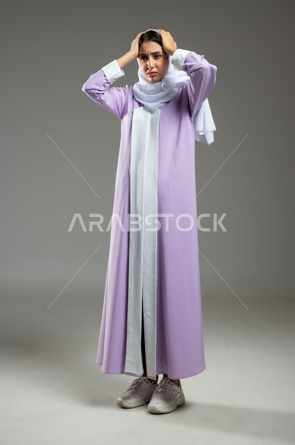 الحاجة للهدوء والراحة، الإحساس بألم الرأس والصداع، المعاناة من آلام الشقيقة، بورتريه لإمرأة عربية خليجية سعودية محجبة ترتدي عباءة وردية تضع يديها على رأسها بايماءات التعب والارهاق، صورة الجسم بالطول الكامل، خلفية رمادية، محتوى حساس، استخدام حساس