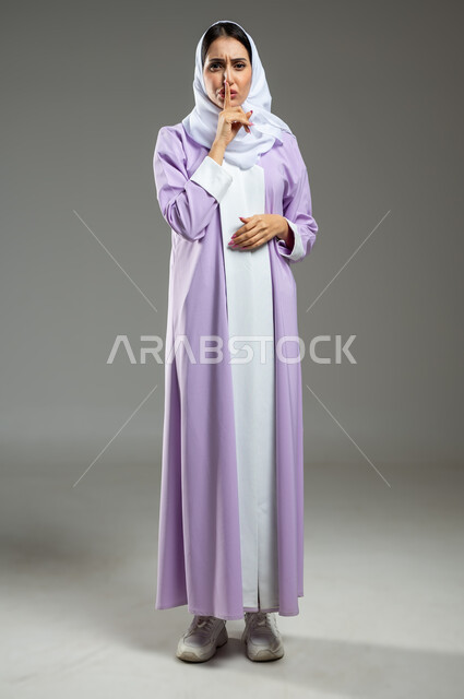 التوقف عن الكلام و الأمر بالصمت و الاستماع، بورتريه لإمرأة عربية سعودية خليجية محجبة ترتدي العباءة تنظر إلى الكاميرا وترفع اصبعها الى فمها، إيماءات يدين تدل على طلب الهدوء و عدم الإزعاج, الجسم بالطول الكامل، خلفية رمادية، استخدام حساس، محتوى حساس