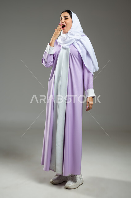Feeling of laziness, lethargy, and the desire for rest, drowsiness, fatigue, and the need to sleep, portrait of a Saudi Arabian Gulf woman wearing the abaya and hijab, placing her hand over her mouth, standing with tired gestures, full-length body, gray background, sensitive use, sensitive content