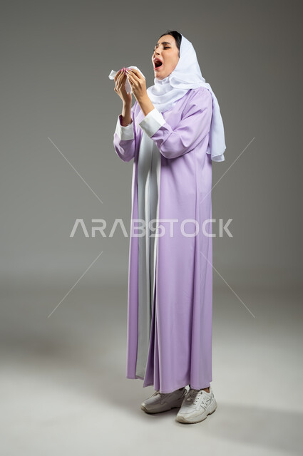 Expressions indicating sneezing and fatigue, feeling tired and sick, catching a cold and flu, using tissues while sneezing, a portrait of a veiled Saudi Arabian Gulf woman wearing an abaya suffering from allergies, full-length body, gray background, sensitive use, sensitive content