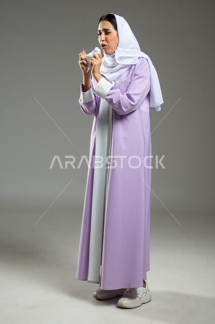 Suffering from cold and flu, expressions indicating sneezing and fatigue, feeling tired and sick, using tissues while sneezing, portrait of a veiled Saudi Gulf Arab woman wearing an abaya suffering from allergies, full-length body, gray background, sensitive use, sensitive content