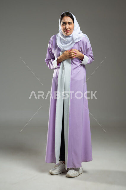 مشاكل صحية صعبة، إيماءات التعب والارهاق والضغوطات النفسية، بورتريه لإمرأة عربية سعودية خليجية محجبة ترتدي العباءة تضع يدها على قلبها تشعر بالحالة سيئة من الوخز والألم، الجسم بالطول الكامل، خلفية رمادية، استخدام حساس، محتوى حساس