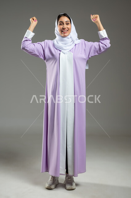 النجاح و تحقيق الاهداف، النظر الى الكاميرا بتعابير الفوز والكسب، ايماءات الفرح للانتهاء من انجاز شيء ما، بورتريه لامرأة عربيه خليجيه سعوديه محجبه ترتدي عباءة عصرية ترفع يديها بحركات التشجيع والحماس، الطول الكامل للجسم، خلفية رمادية، استخدام حساس، محتوى حساس