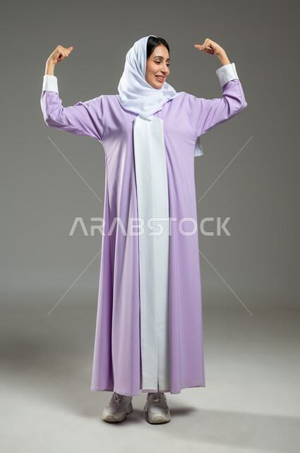 ايماءات الفرح للانتهاء من انجاز شيء ما، النجاح و تحقيق الاهداف، تعابير الفوز والكسب، بورتريه لامرأة عربيه خليجيه سعوديه محجبه ترتدي عباءة عصرية ترفع يديها بحركات التشجيع والحماس، الطول الكامل للجسم، خلفية رمادية، استخدام حساس، محتوى حساس