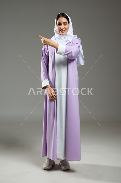 Expressions of certainty and locating a specific place, looking at the camera with gestures of happiness and pleasure, portrait of a veiled Saudi Gulf Arab woman, smiling, wearing a modern abaya, pointing with her index finger at something, full-length body, gray background, sensitive content, sensitive use