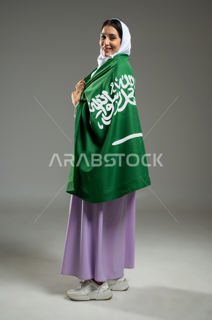 الأعياد والمناسبات الوطنية، بورتريه من الجانب لشابة عربية خليجية سعودية محجبة ترتدي علم السعودية بإيماءات تدل على السعادة، إحياء ذكرى العيد الوطني السعودي 23 سبتمبر، يوم العلم 11 مارس، صورة الجسم بالطول الكامل، خلفية رمادية، محتوى حساس، استخدام حساس